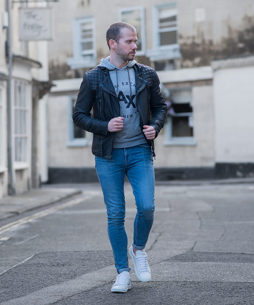 Black biker leather jacket, gray hoodie, blue jeans, and white sneakers outfit