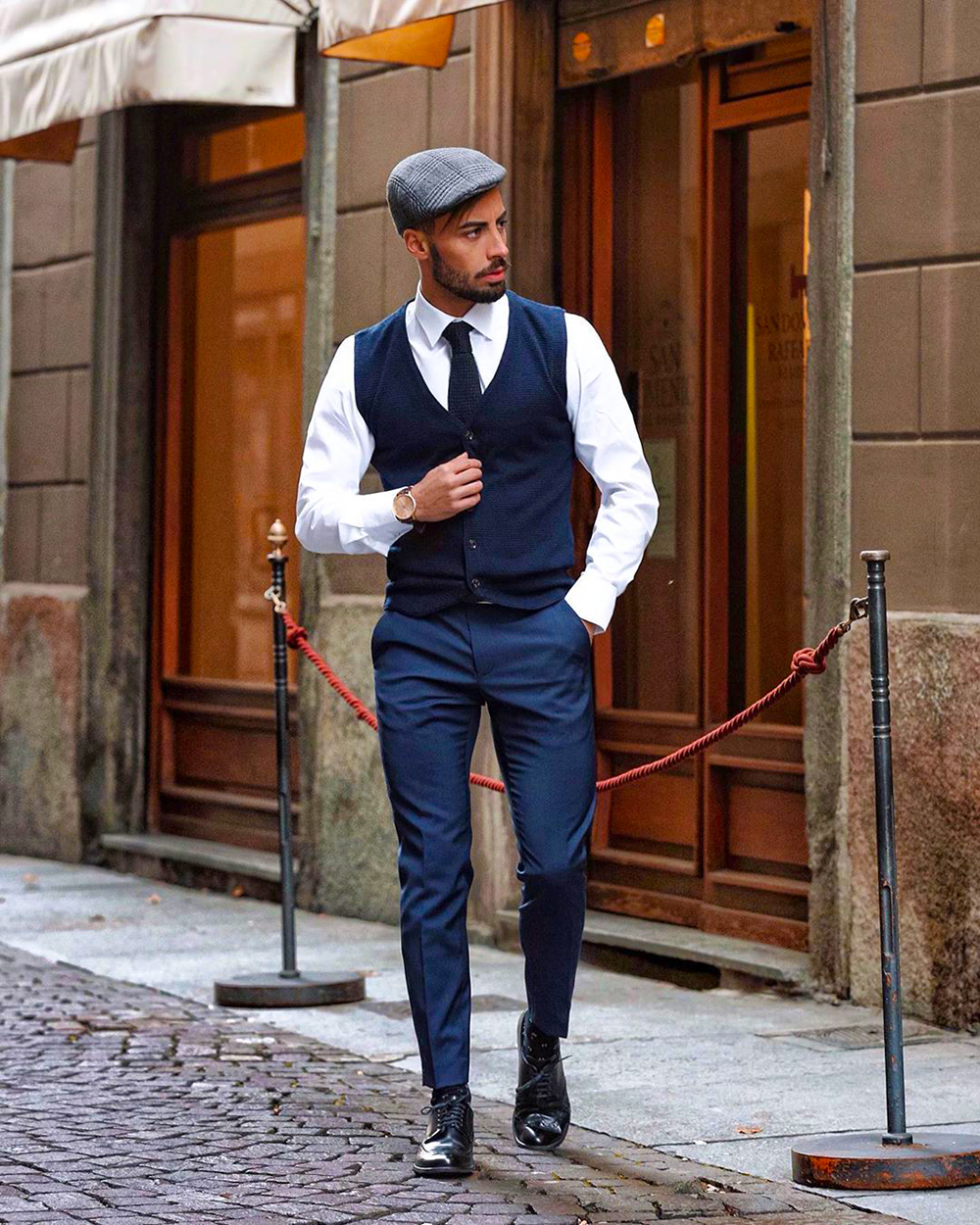 Navy waistcoat, white dress shirt, navy tie, navy dress pants, gray cap, and black derby shoes outfit