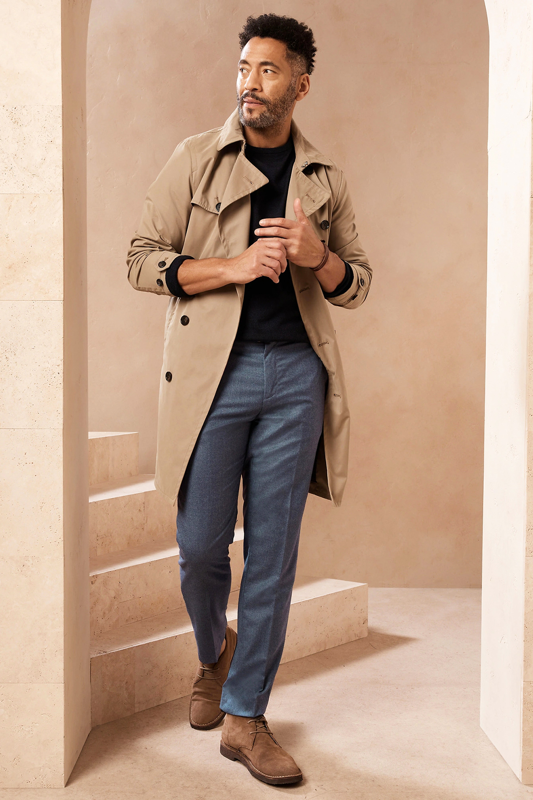 Tan trench coat, black crew neck sweater, blue dress pants, and light brown chukka boots outfit
