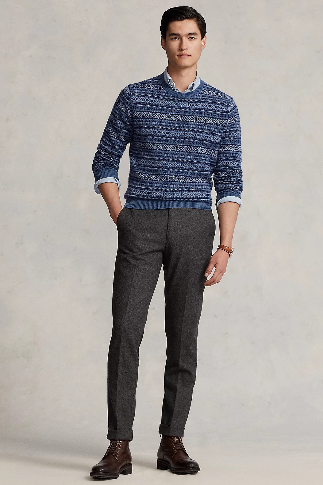 Wearing a blue fair isle wool sweater, light blue dress shirt, charcoal gray dress pants, and dark brown lace-up boots